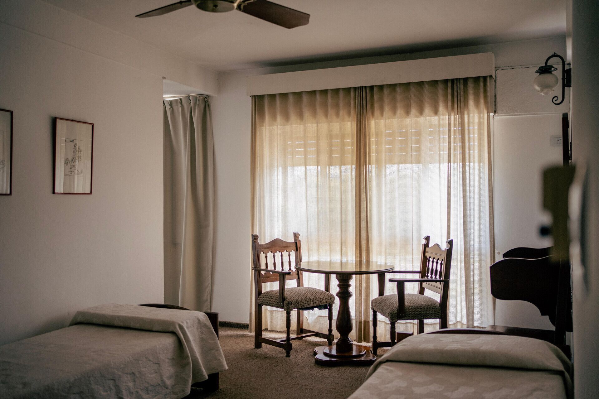 A hotel room with two single beds, a small round table with chairs, a ceiling fan, and a large window with sheer curtains.