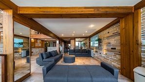 Un hall d’entrée spacieux, avec un grand canapé gris, un canapé plus petit et une table basse. Le hall est agrémenté de poutres en bois, de murs en pierre et d’un grand écran de télévision.