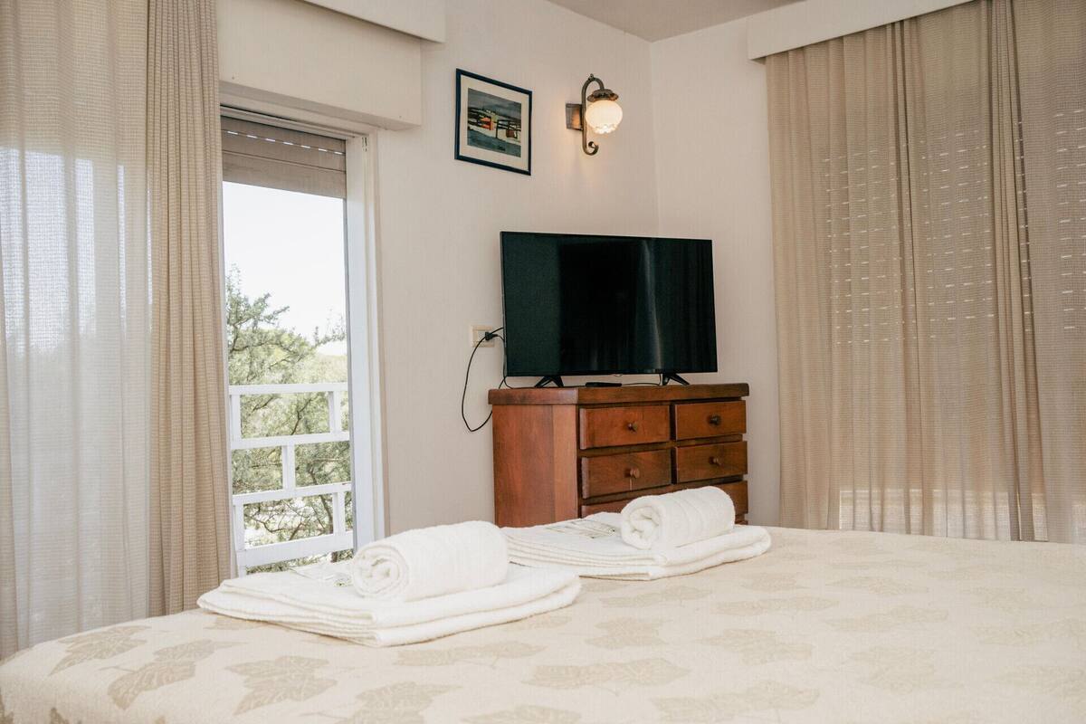 Een slaapkamer met een bed, een houten dressoir, een televisietoestel en een raam met gordijnen.