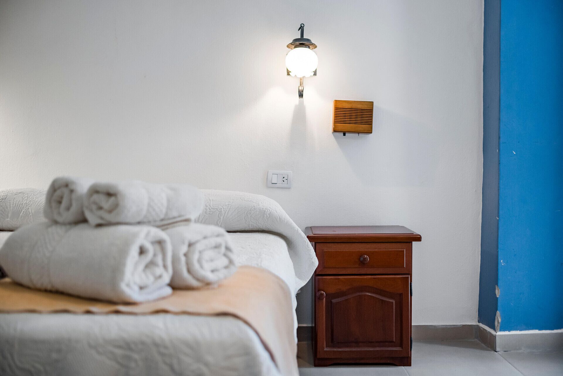Una cama bien hecha con toallas blancas enrolladas, una mesita de noche con cajón, una lámpara de pared y un panel de madera pequeño.