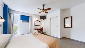 Une chambre d’hôtel comprenant un lit, un ventilateur de plafond, une télévision, un bureau avec une chaise et une fenêtre avec des rideaux.