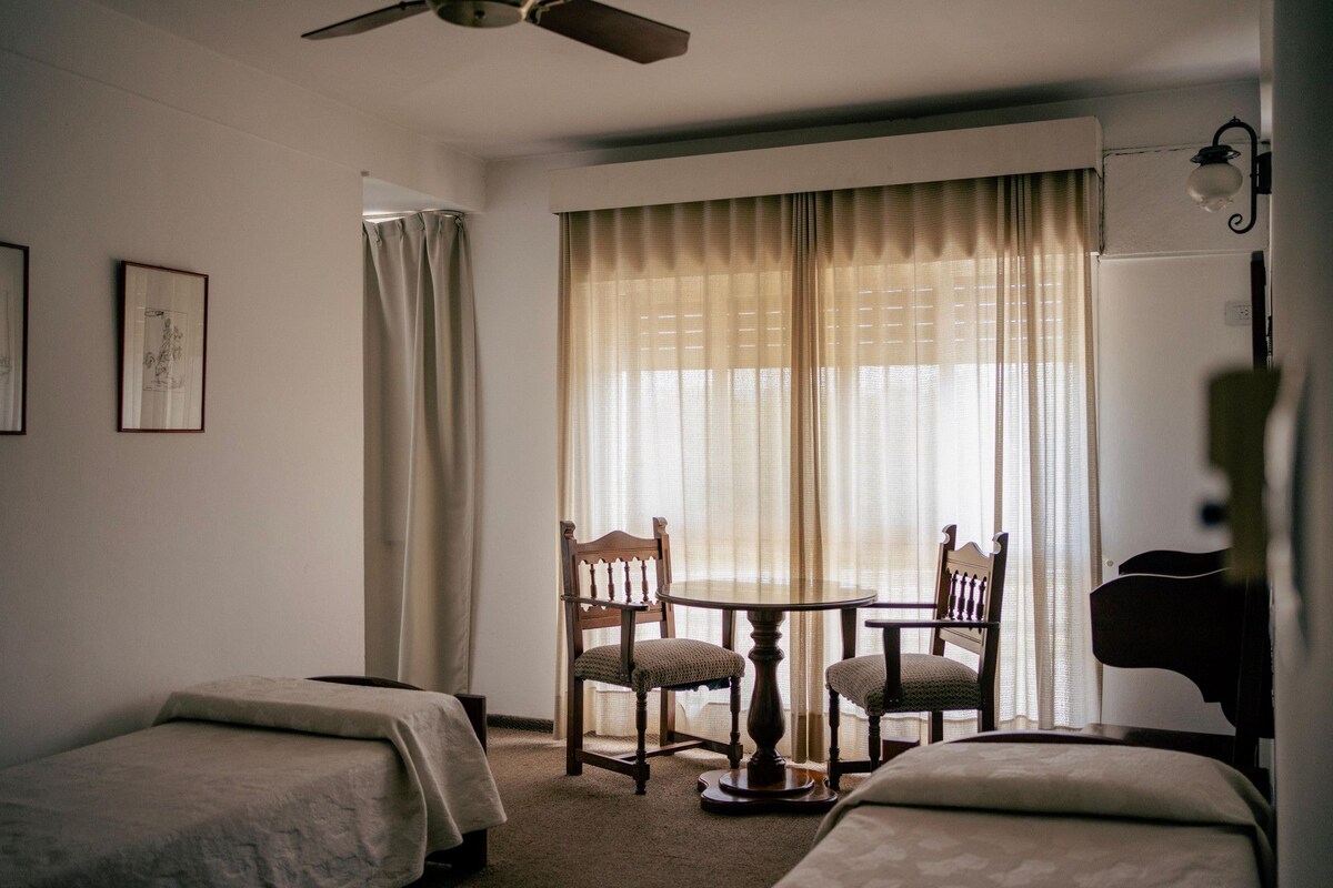 Una habitación de hotel con dos camas individuales, una mesita redonda con sillas, un ventilador de techo y un ventanal con cortinas traslúcidas.