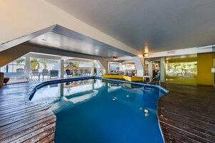 Una piscina interior con cubierta de madera, agua azul cristalina y una zona de estar con sillas y una mesa.