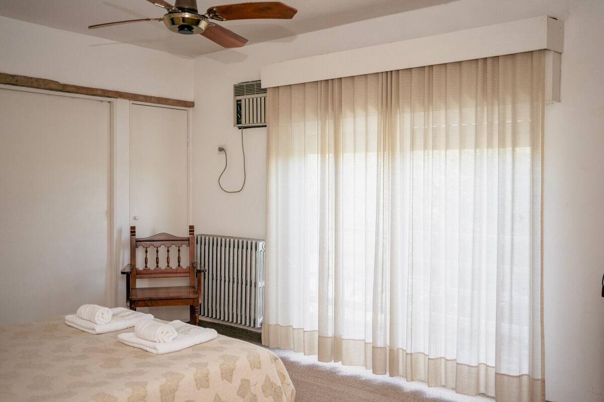 Une chambre à coucher avec un lit, une tête de lit en bois, un radiateur, un ventilateur de plafond et des rideaux en voile.