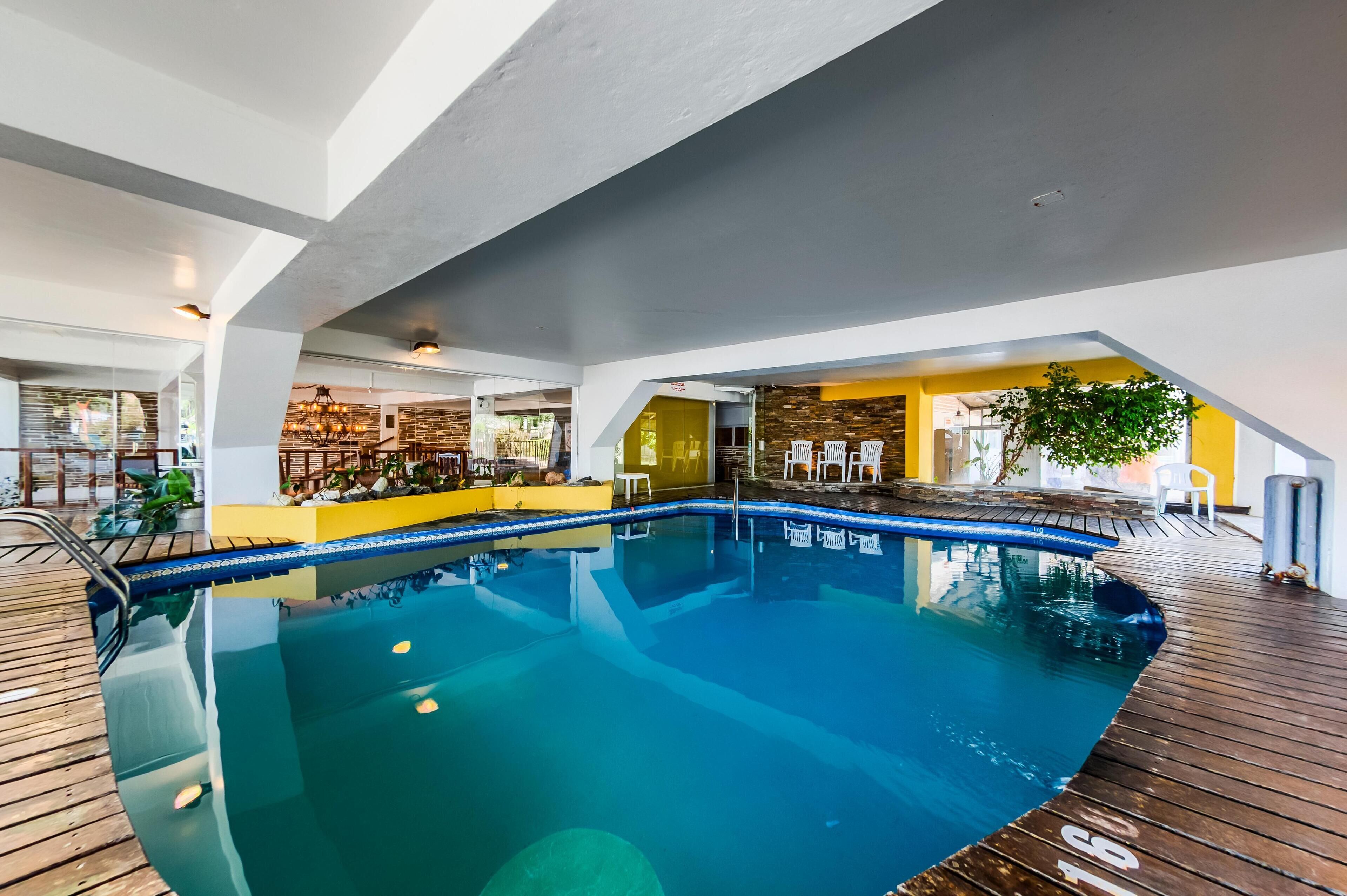 Una piscina interior con plataforma de madera, rodeada de paredes amarillas y blancas, y un área de descanso con sillas y una planta.