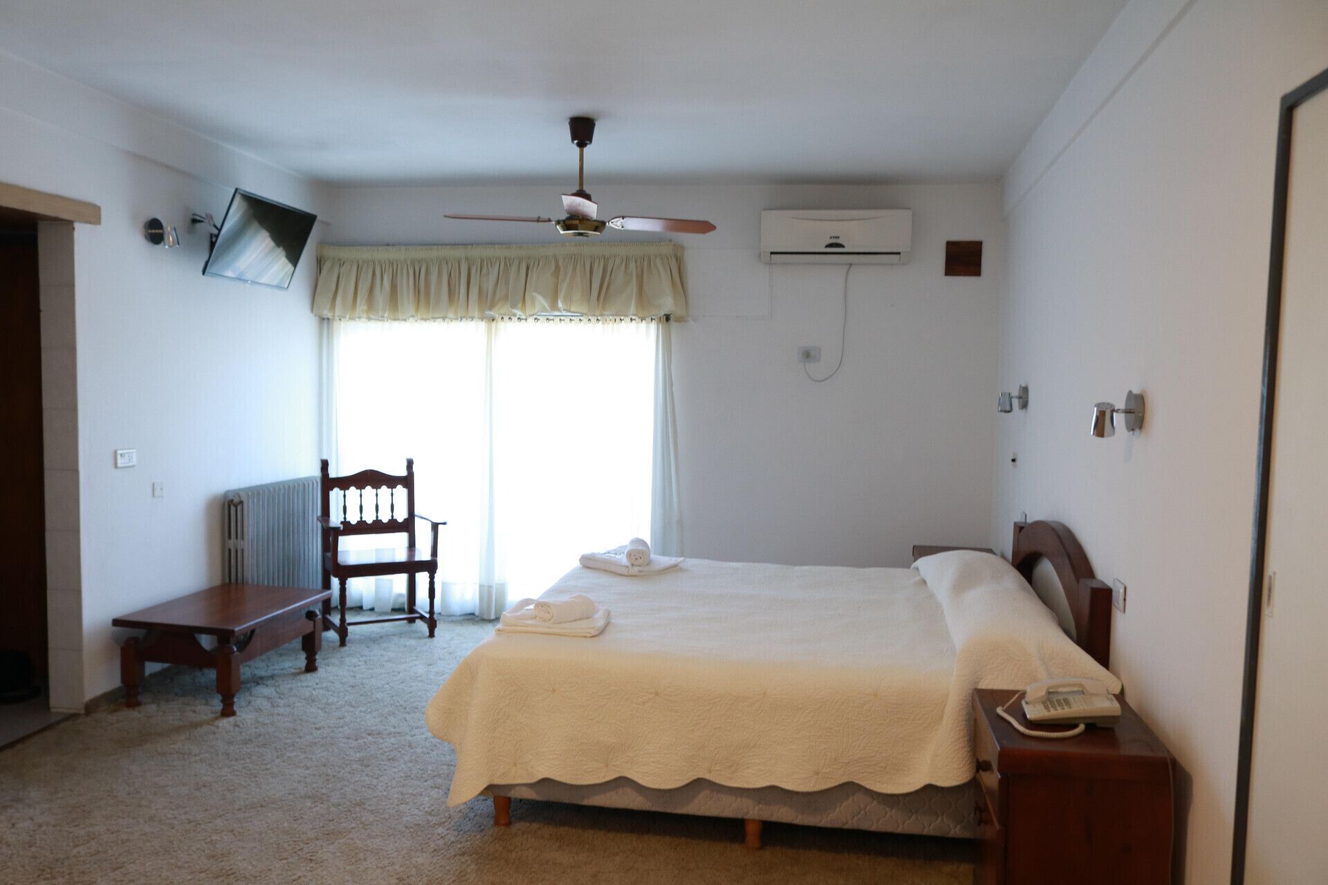 Een slaapkamer met een bed, een stoel, een bureau, een telefoon en een plafondventilator.