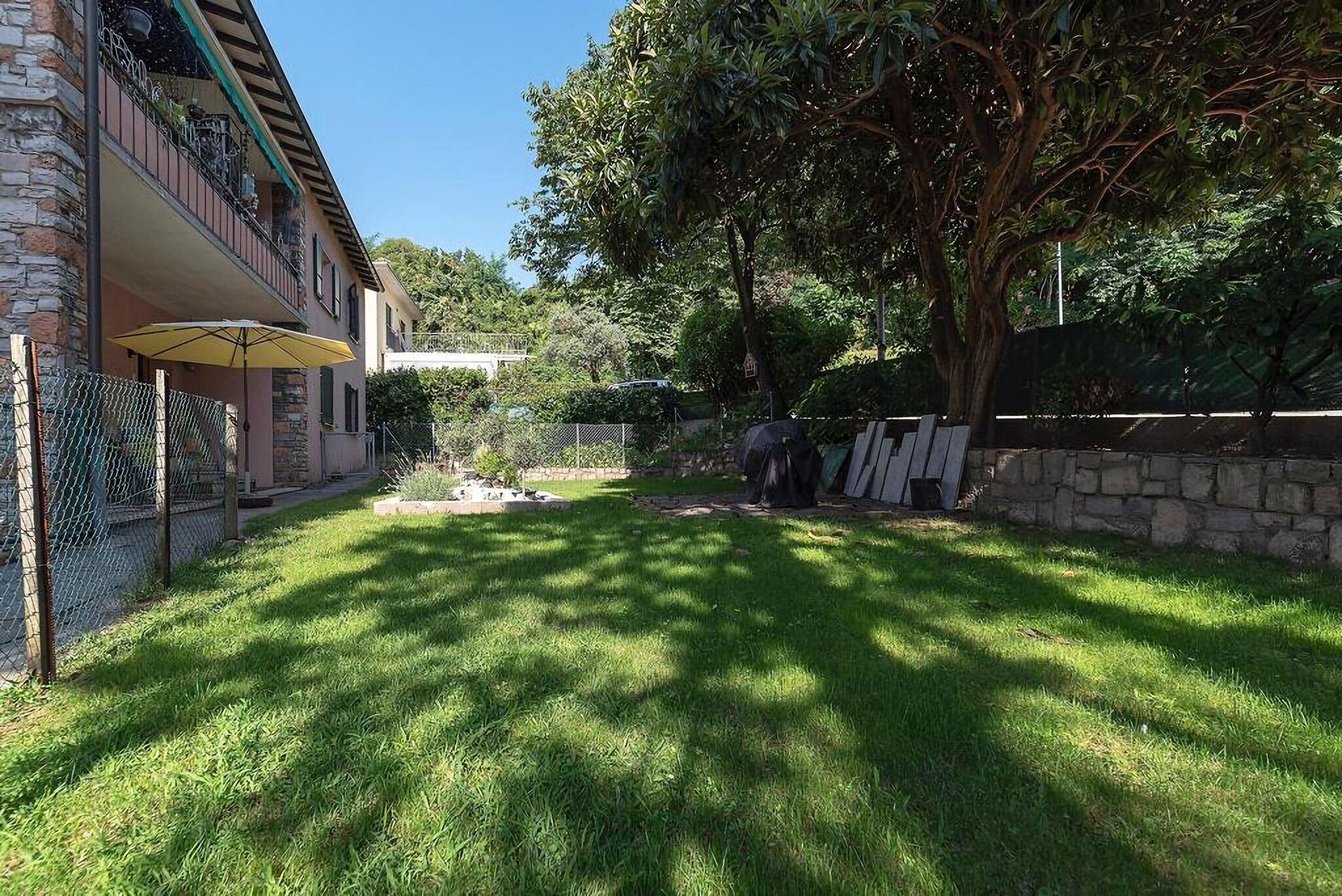 A well-maintained garden with a stone wall, a yellow umbrella, and a large tree providing shade.