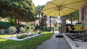 Zona patio con ombrellone giallo, vialetto in pietra, posti a sedere all'aperto e vegetazione.