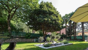 Un giardino curato con diversi alberi e arbusti, un muro in pietra e una zona pavimentata con un ombrellone giallo.