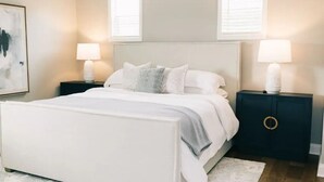 A bedroom with a white bed, two bedside tables with lamps, a dark nightstand with a circular design, and a painting on the wall. - Cozy, Clean Home for Family or Friends (Bowling Green)