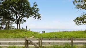 Une clôture en bois avec un portail donnant sur une zone herbeuse, parsemée d’arbres, et offrant une vue sur la mer.