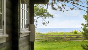 Vue depuis une maison en bois dotée d’une fenêtre, donnant sur une zone herbeuse bordée d’une clôture en bois, avec une étendue d’eau visible au loin.