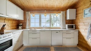Une cuisine compacte aux murs en bois, avec des meubles blancs, un four encastré, un four à micro-ondes et une fenêtre agrémentée de rideaux.