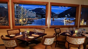 A dining area with tables set for four, large windows offering a view of a snowy mountain and a town below. - Cozy Keystone Lodge Stay with Breathtaking Views (Keystone)