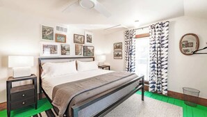 Ein Schlafzimmer mit Bett, Nachttischen, Deckenventilator, gerahmten Kunstwerken an der Wand und gemusterten Vorhängen.