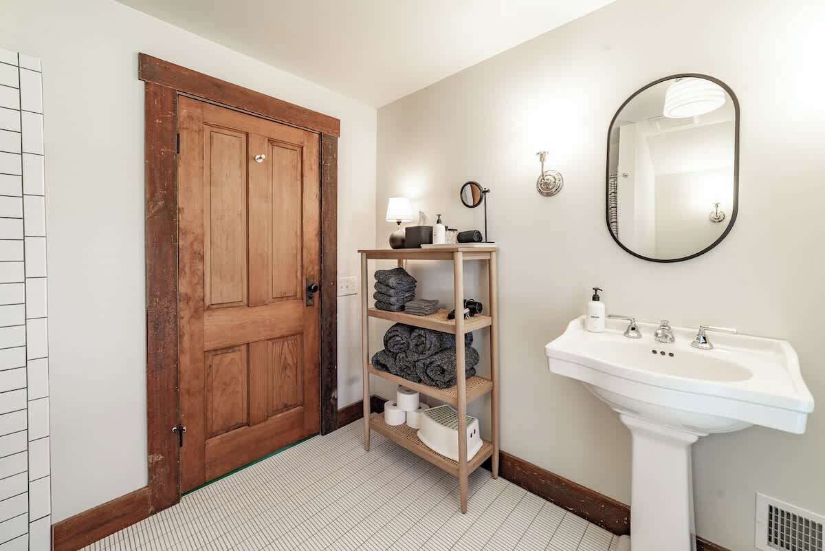 A bathroom with a wooden door, a white sink with a mirror, and a shelf with towels and toiletries.