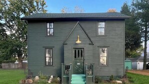 Ein zweistöckiges Haus mit grüner Fassade, einer Veranda mit Stufen und einem kleinen Garten.