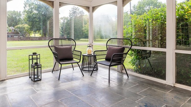 Une terrasse couverte avec deux chaises et une petite table, de grandes fenêtres offrant une vue sur un jardin et des arbres.