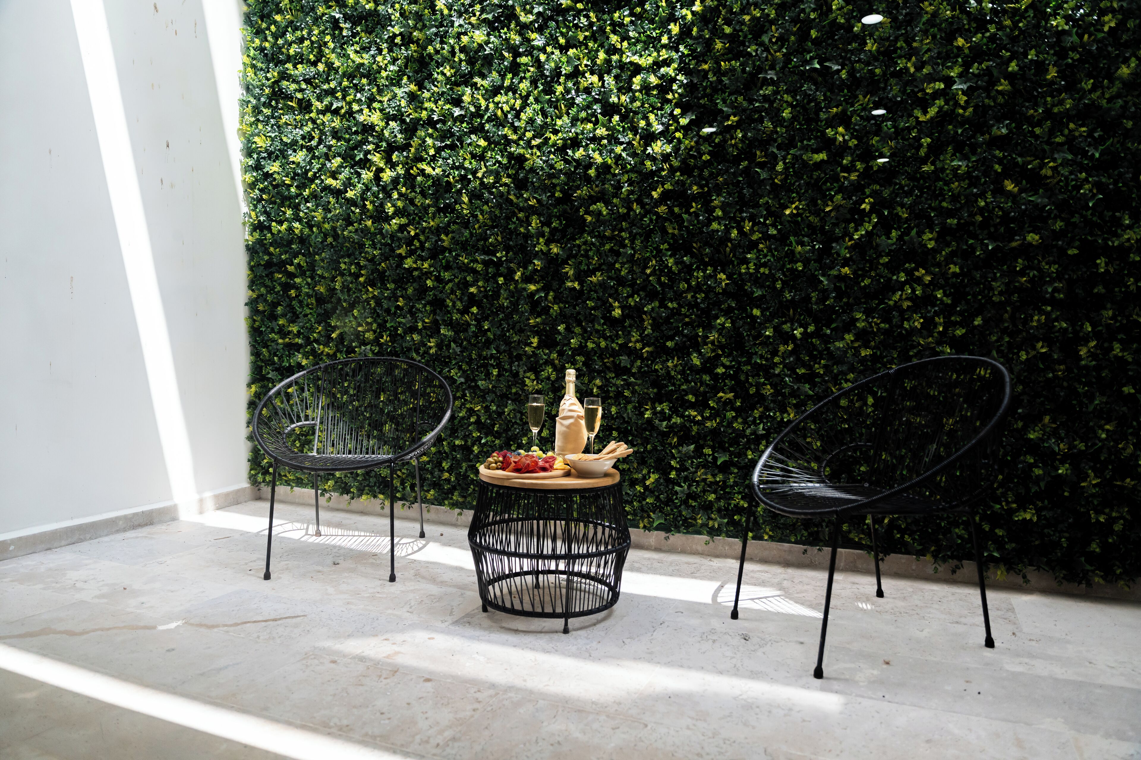 A small round table with a bottle and glasses, two chairs, and a green hedge wall.