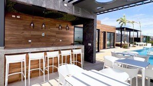 Ein moderner Außenbereich mit Bar, Pool, weißer Möbel und einer Holzwand mit hängenden Lampen.