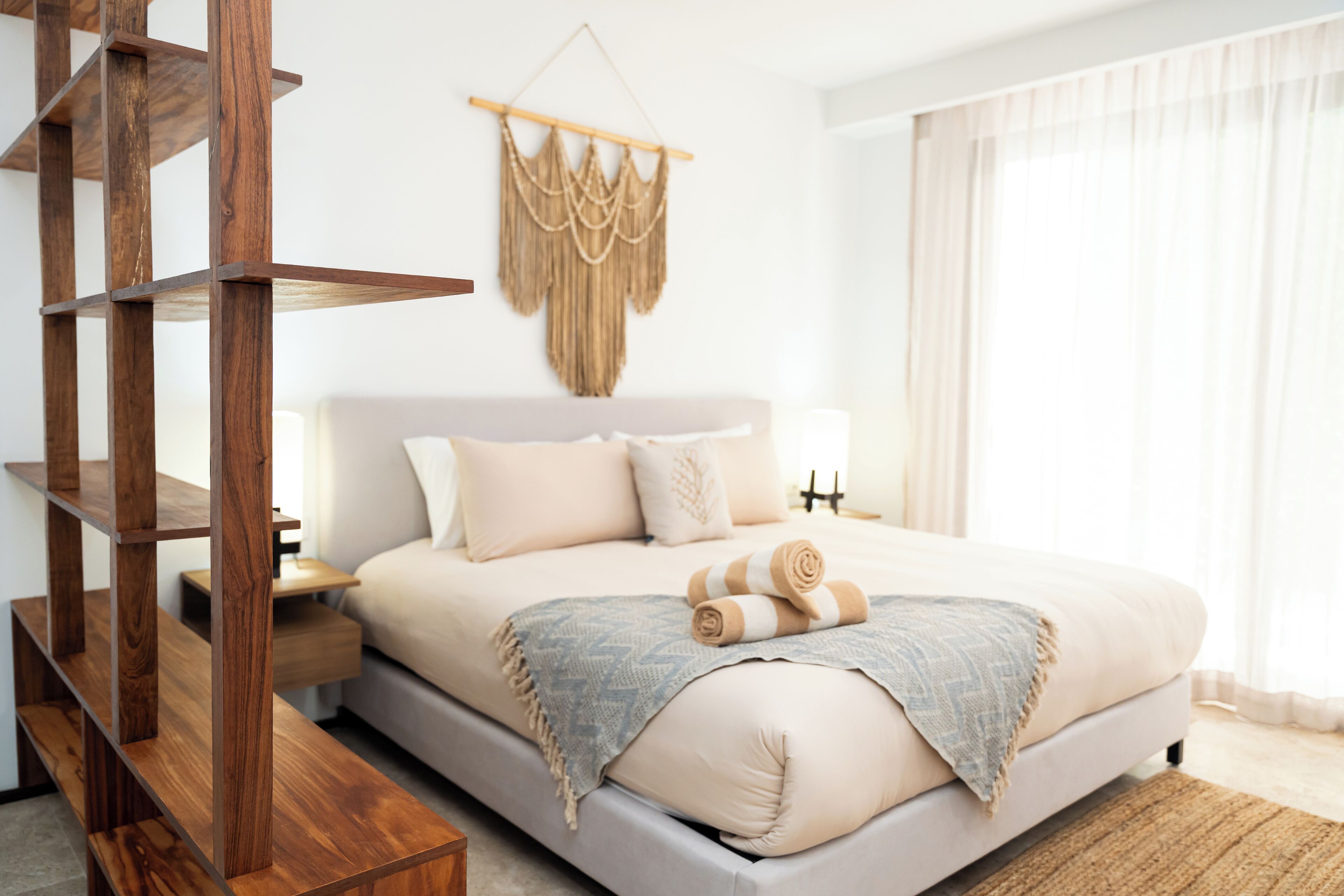 Een moderne slaapkamer met een groot bed, houten nachtkastjes, een macramé wanddecoratie en uitzicht op het groen door dunne gordijnen.