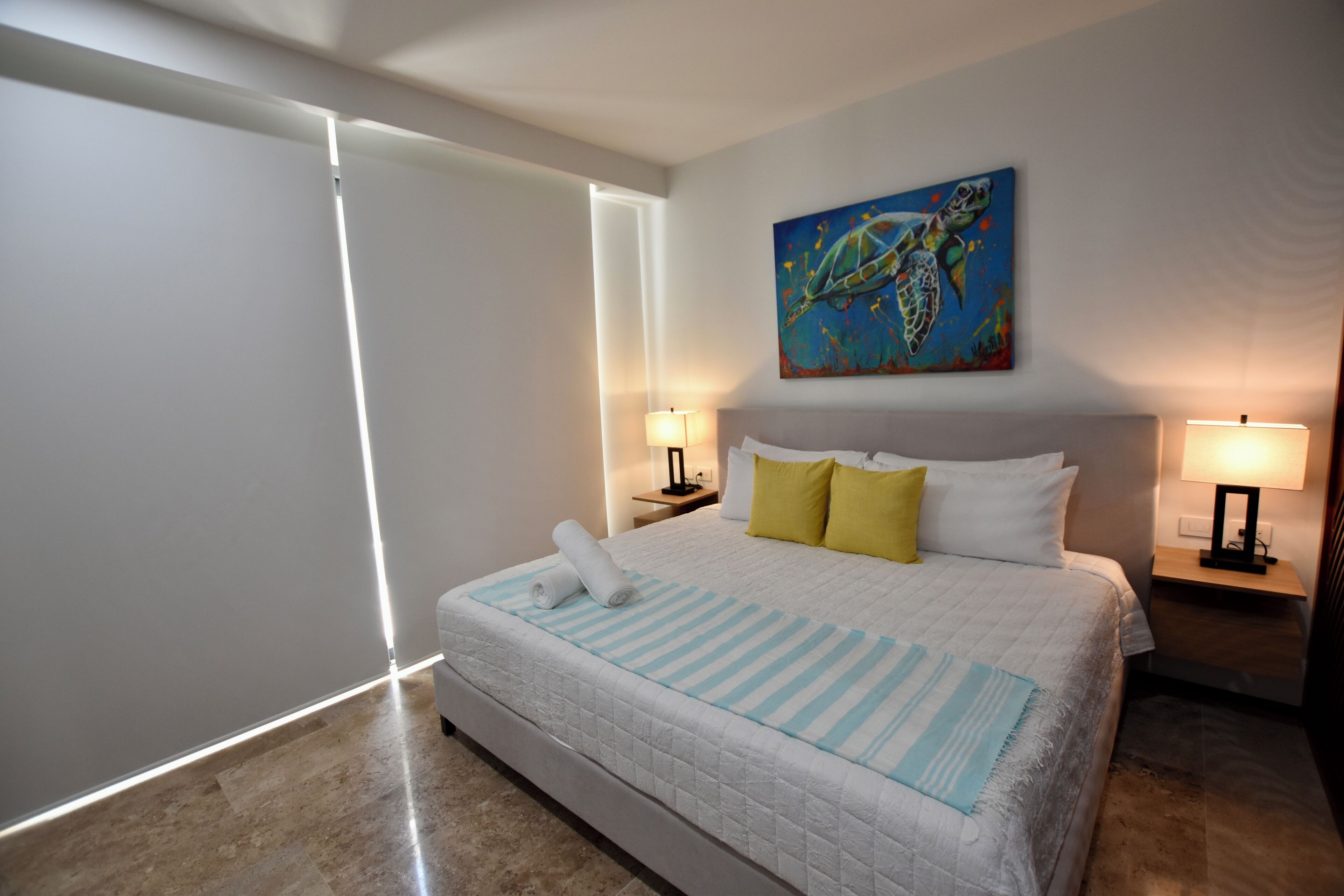 Une chambre d’hôtel avec un grand lit, deux tables de chevet avec des lampes, un tableau au mur et une fenêtre avec des stores.