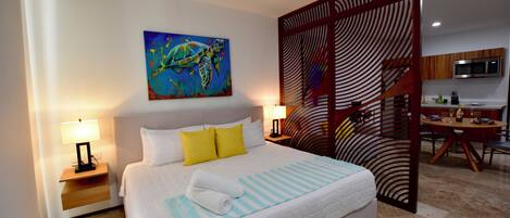 A hotel room with a bed, two bedside lamps, a painting of a sea turtle, a wooden partition, a desk, and a chair.