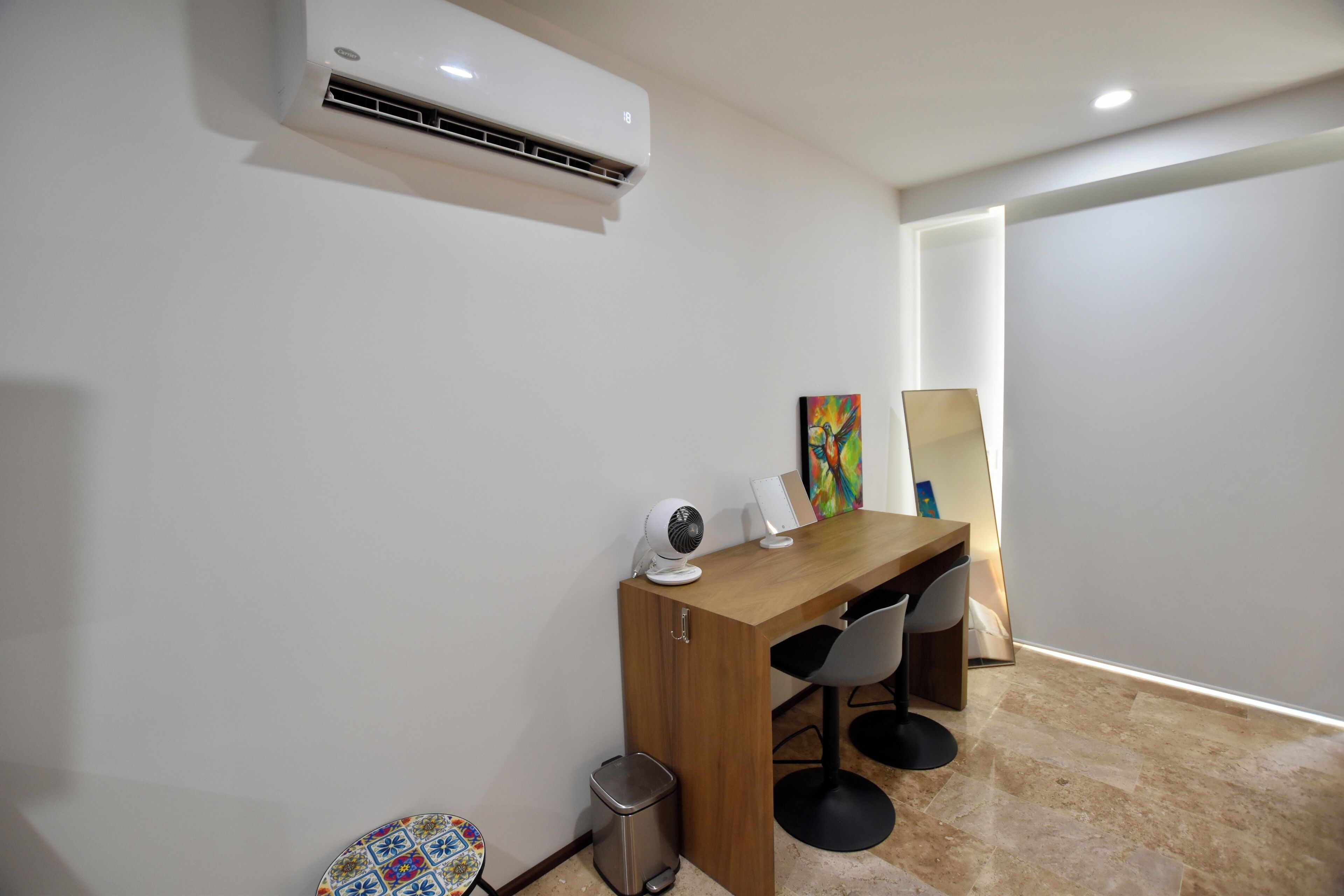 Un espacio de oficina moderno con un escritorio de madera, dos sillas, un aire acondicionado blanco, un cesto de basura y un cuadro de colores en la pared.