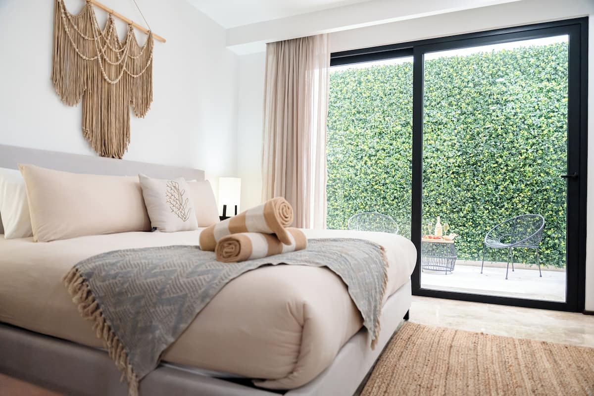 Een slaapkamer met een groot bed, een macramé wanddecoratie, uitzicht op groen via een schuifdeur en een klein terras.
