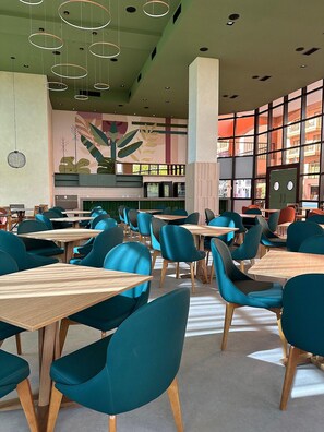 A modern restaurant interior with teal chairs and wooden tables, large windows with a view of buildings, and hanging circular light fixtures. - Jurerê Beach Village é Destino Floripa (Florianópolis)