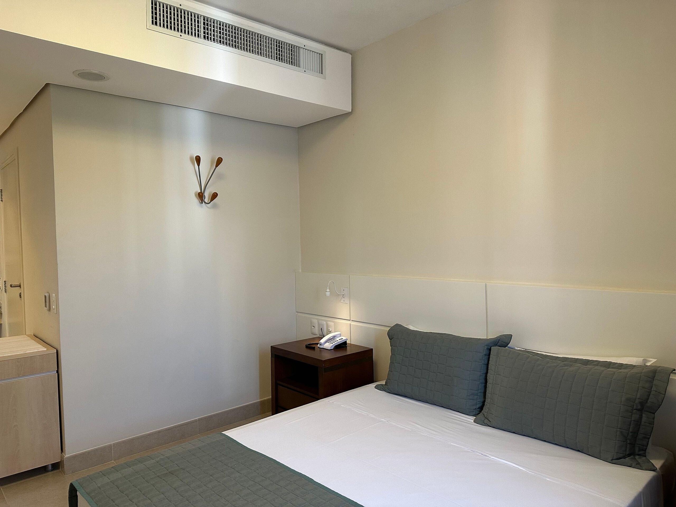 A hotel room with a bed, a bedside table with a phone, a wall-mounted heater, and a door leading to a bathroom.