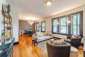 Een moderne woonkamer met een bank, fauteuils, een salontafel en een eethoek met een tafel en stoelen. Grote ramen met jaloezieën, een boekenkast en een kroonluchter.