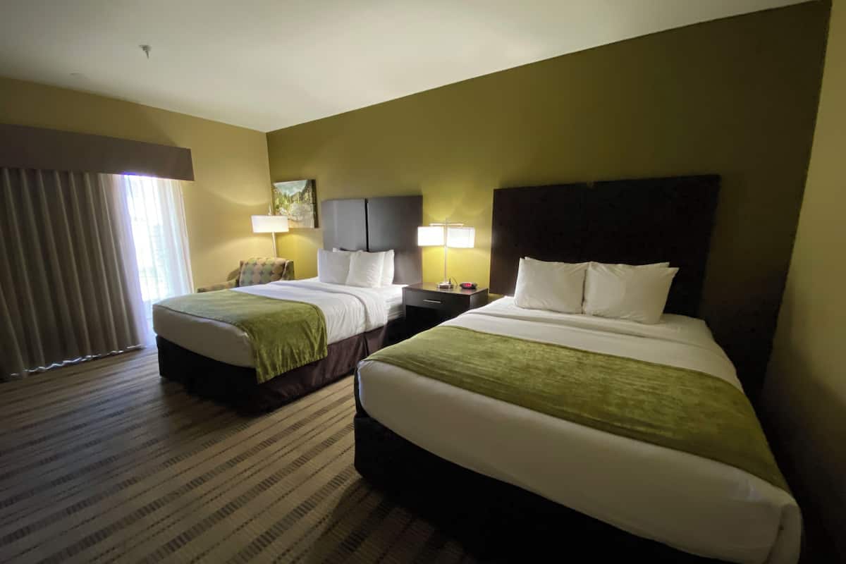 A hotel room with two beds, a large window with curtains, a headboard, and a bedside table with a lamp.