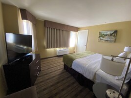Habitación de hotel con televisión de pantalla plana, una cama con manta verde, lámpara de noche, un cuadro en la pared y una ventana con cortinas.