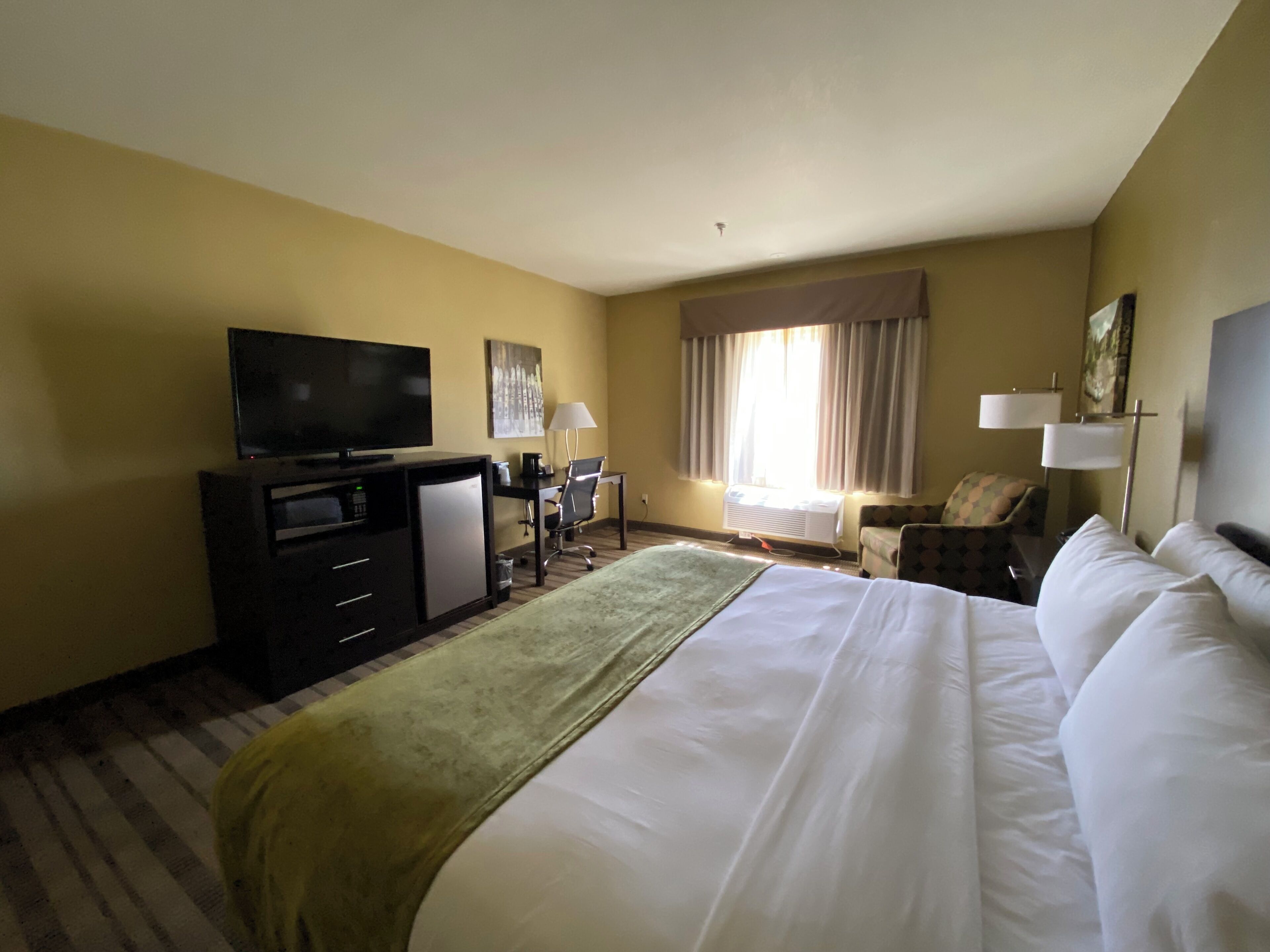 A hotel room with a large bed, a flat-screen TV, a desk with a chair, a small table, and a window with curtains.