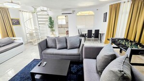 A modern living room with a grey sofa, a coffee table, and a foosball table. There is a staircase with a railing, a dining area with a table and chairs, and a kitchen in the background. - Linkzup Playa Laguna Villa (Sosúa)