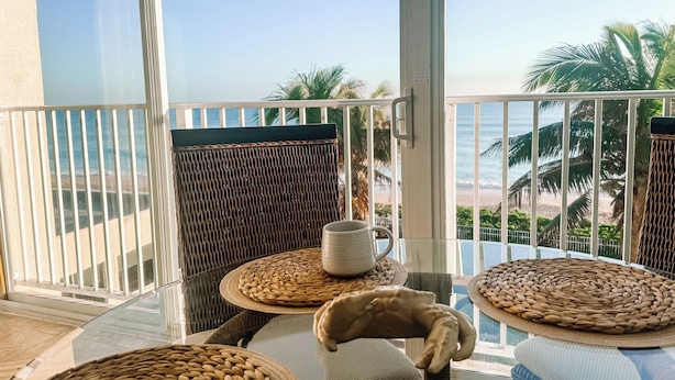 Un balcón con mobiliario de mimbre, una taza blanca y vista a la playa y palmeras.
