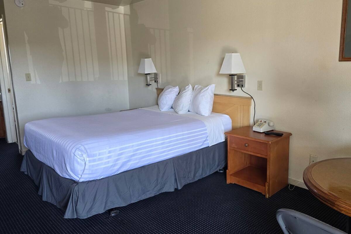 Une chambre d’hôtel comprenant un lit, deux lampes de chevet, un téléphone sur la table de chevet, une table de nuit en bois, une table ronde et une chaise.