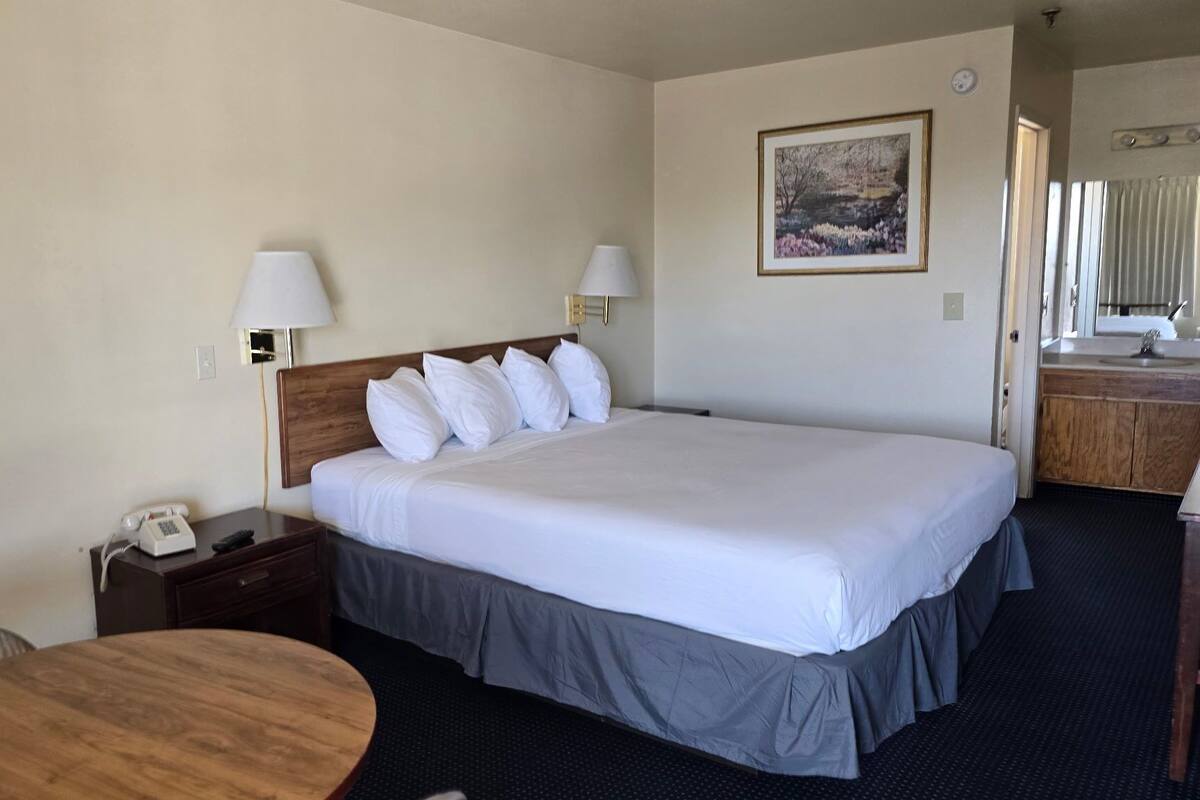 Une chambre d’hôtel avec un grand lit, deux lampes de chevet, un téléphone sur la table de chevet, une table en bois et une salle de bain visible à travers une porte ouverte.
