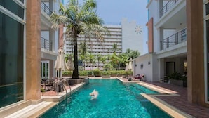 A swimming pool with a person swimming, surrounded by palm trees and a building in the background. - 6-17 - 2 Bdr Condo With Balcony and Shared Pool (Patong)