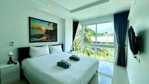 A hotel room with a large bed, a painting of a beach scene, a TV, and a window with a view of greenery. - 6-17 - 2 Bdr Condo With Balcony and Shared Pool (Patong)