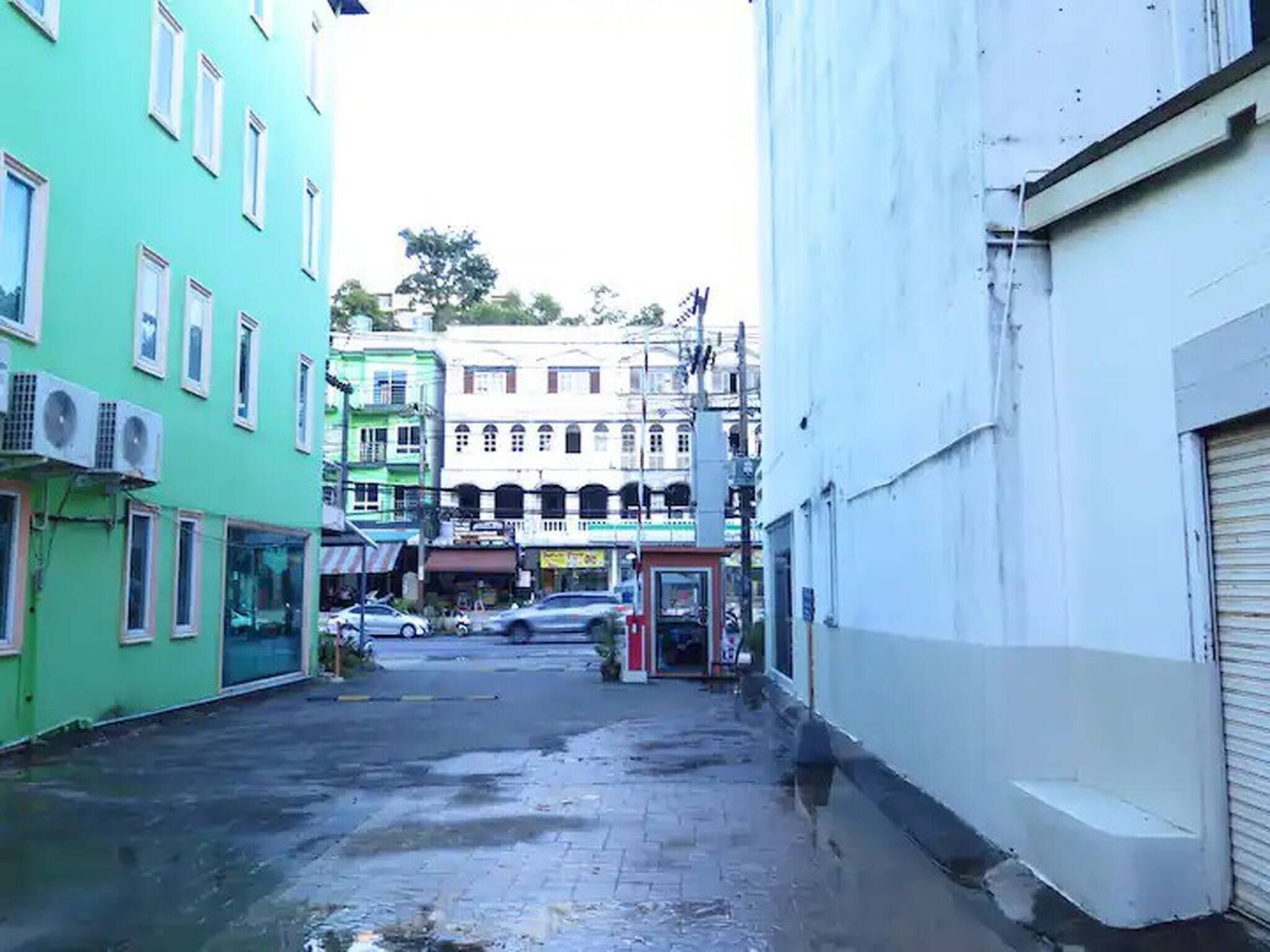 A narrow alleyway with buildings on either side, some with air conditioning units. There are parked cars and a visible storefront in the background.