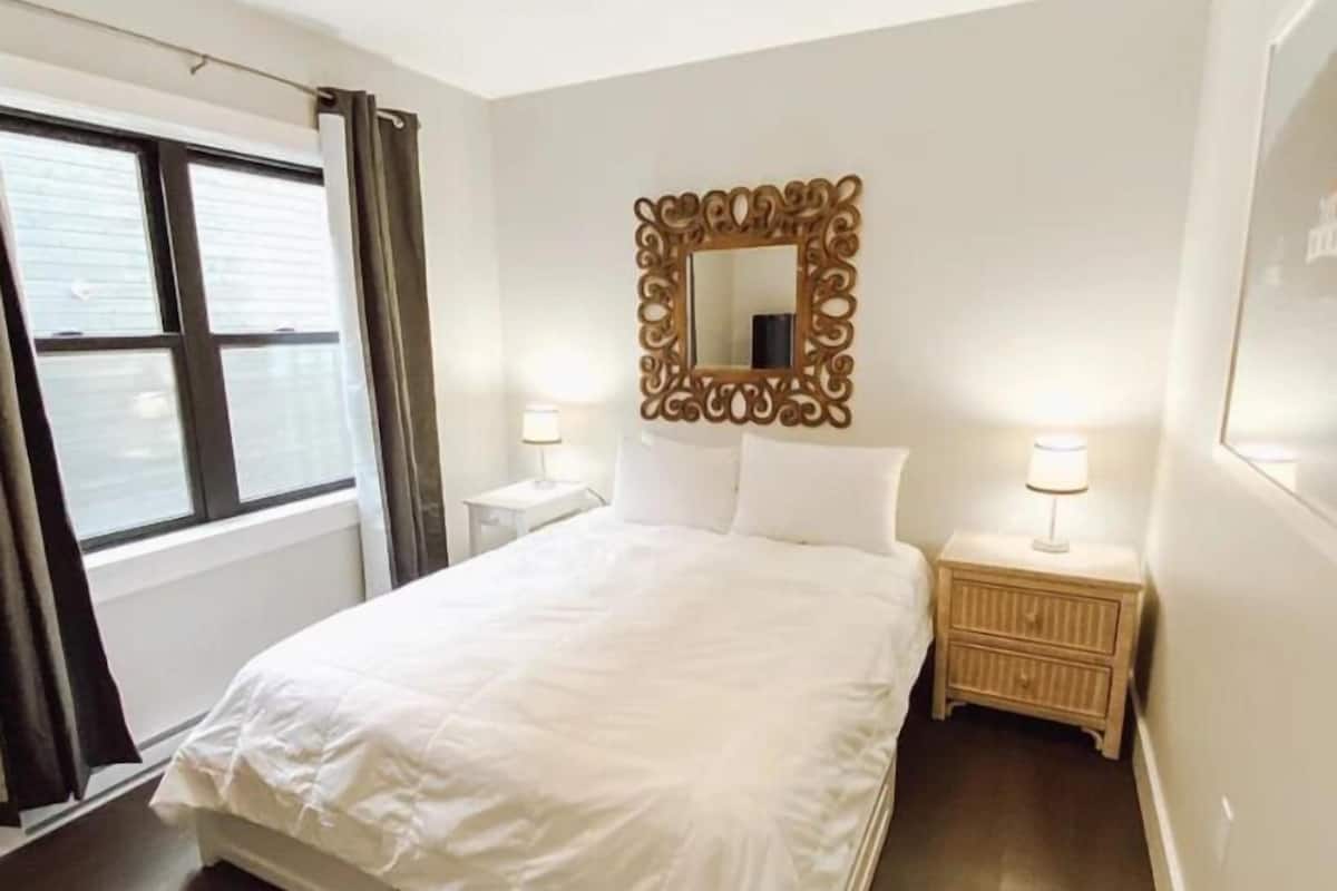 Ein Hotelzimmer mit einem großen Bett, einem Deckenventilator, einem Fenster mit Vorhängen, einem dekorativen Spiegel und Nachttischlampen.