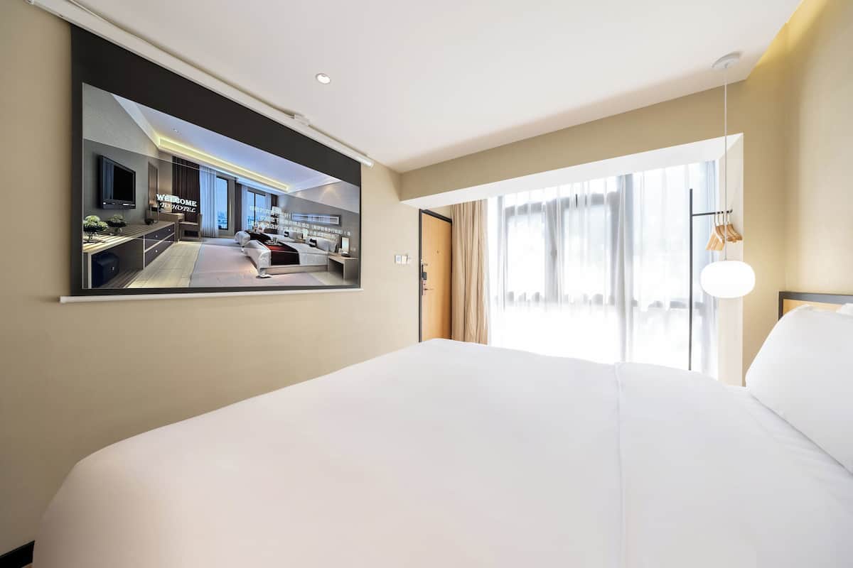 Une chambre d’hôtel avec un grand lit, un téléviseur fixé au mur, une grande fenêtre avec des rideaux et une vue sur le hall de l’hôtel.