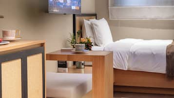 A hotel room with a bed, a small wooden table with a cup and saucer, a television mounted on the wall, and a window with curtains.
