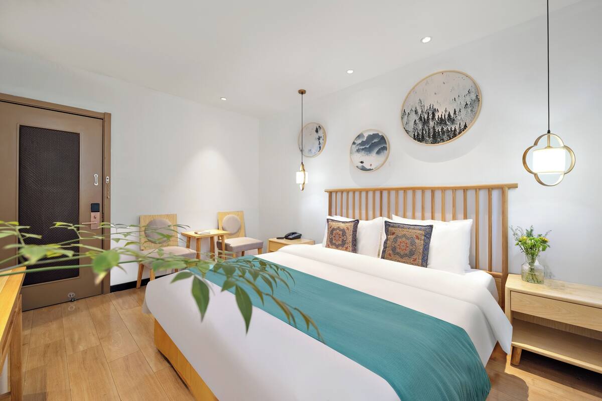 Une chambre d’hôtel moderne avec un grand lit, une table de chevet en bois, un bureau et une chaise. Des œuvres d’art ornent les murs, et un miroir est accroché au mur.