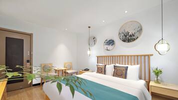 Une chambre d’hôtel moderne avec un grand lit, une table de chevet en bois, un bureau et une chaise. Des œuvres d’art ornent les murs, et un miroir est accroché au mur.