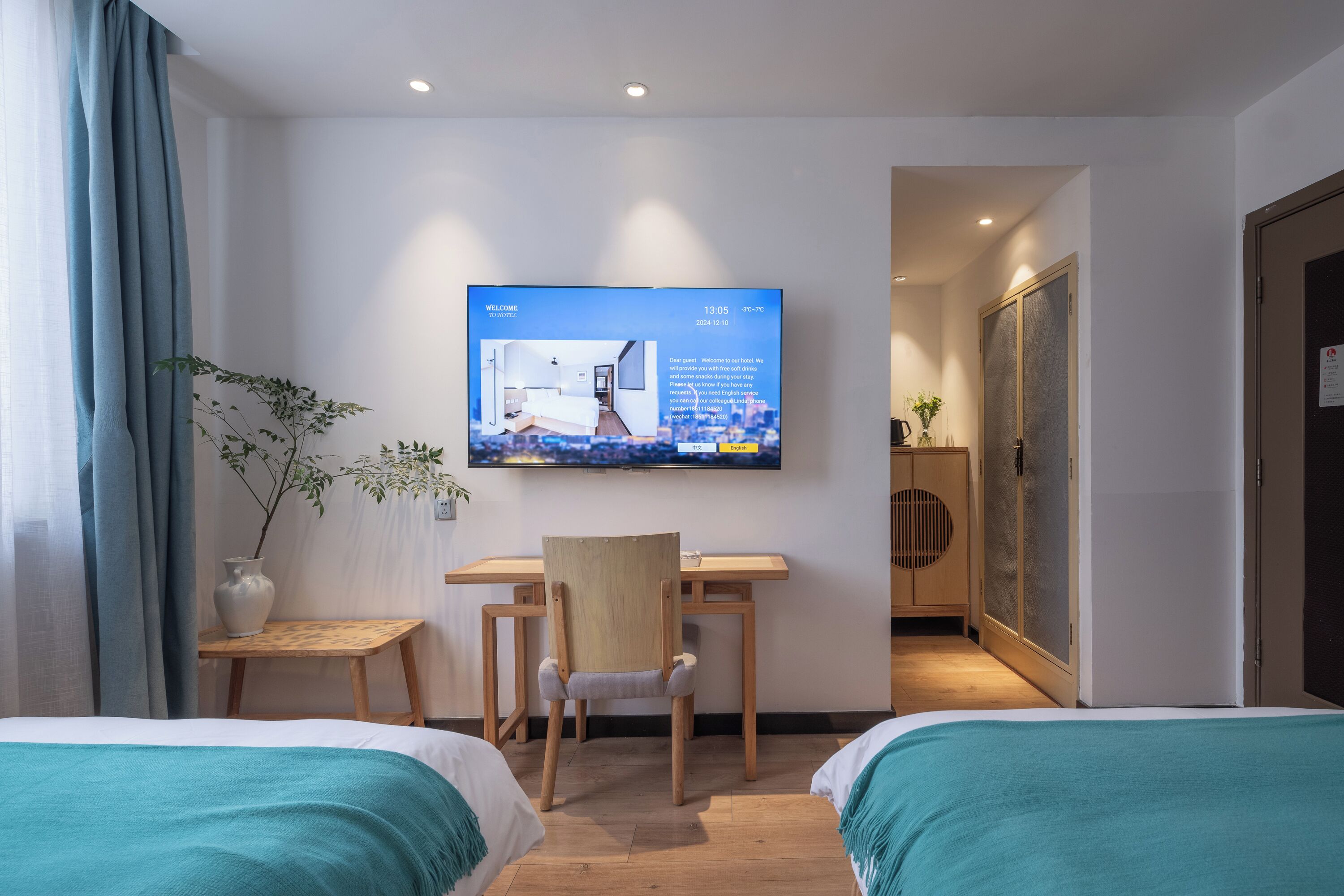 A hotel room with a bed, a desk with a chair, a TV on the wall, and a plant.
