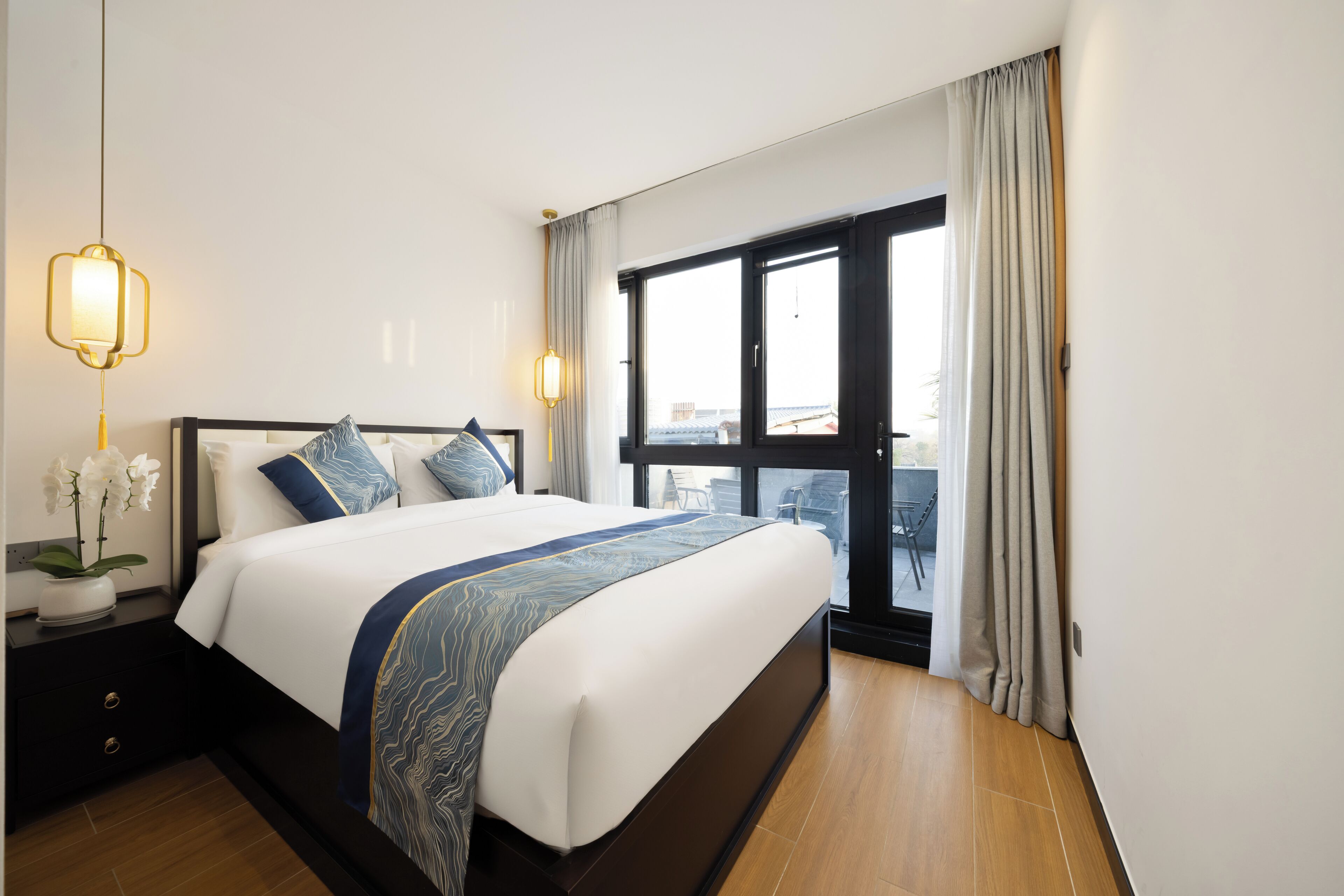 A hotel room with a large bed, a nightstand with a lamp, a window with curtains, and a view of the outside.