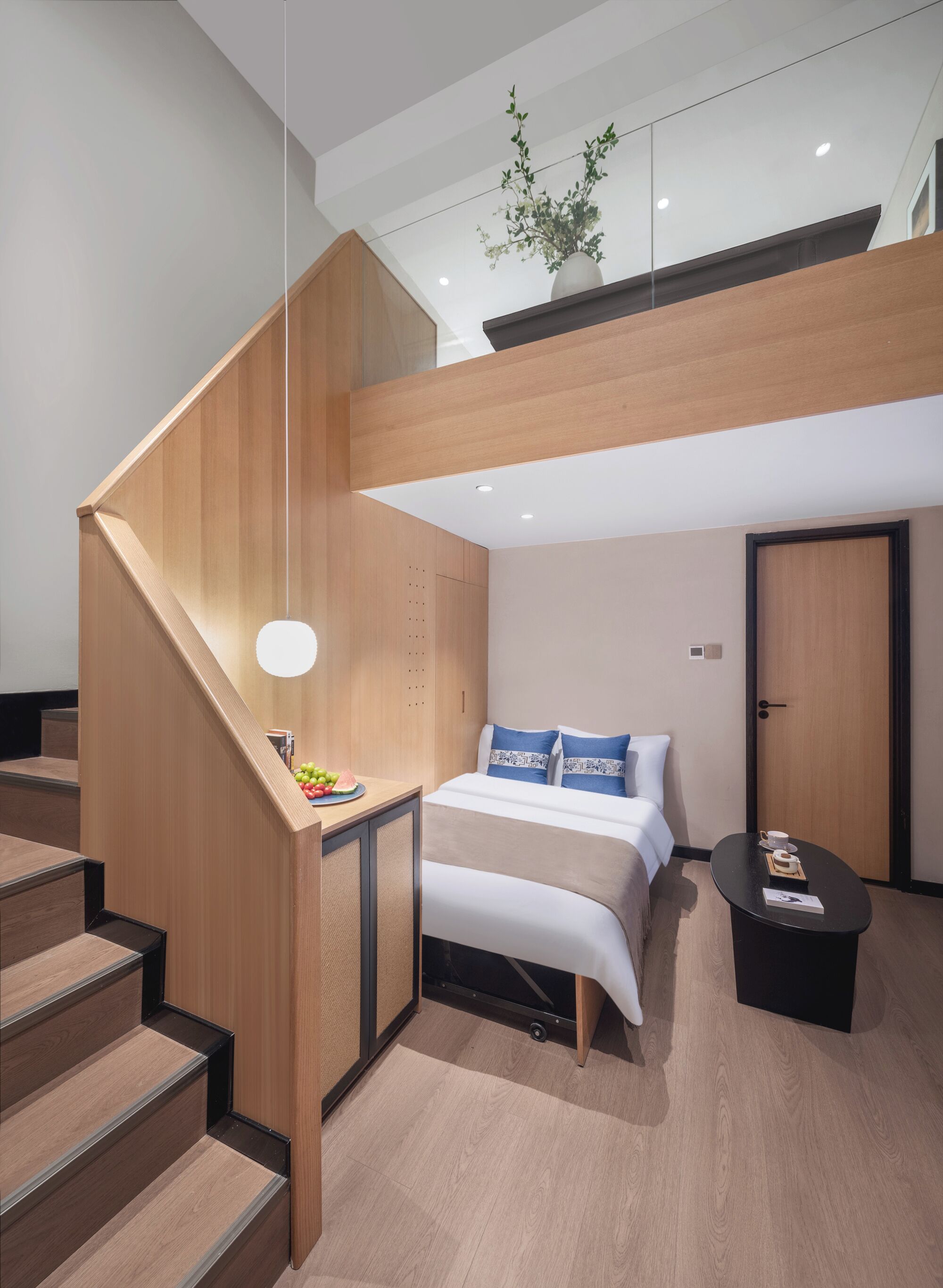 A modern hotel room with a wooden staircase, a bed with blue and white bedding, a small table with a cup and saucer, and a potted plant on the ceiling.